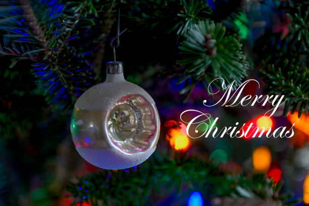 A circa 1950 Christmas ornament on a pine limb with multicolored lights in the background ad the words \