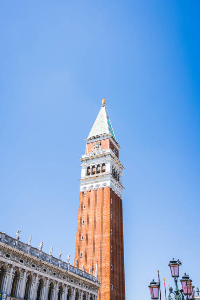 A low-angle view of the iconic Campanile of St. Mark’s Basilica in Venice, Italy. This brick bell tower stands tall against a bright blue sky, showcasing its timeless architectural elegance and historical significance