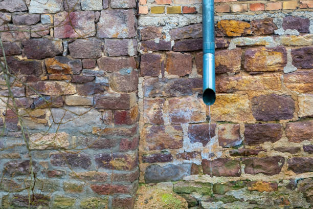 istock Water draining from a metal downspout against an old stone wall covered in moss and historical texture 2192539829