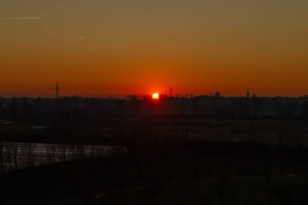 This image captures a serene sunset over the outskirts of Milan, Italy. The fiery red sun is low on the horizon, casting a warm orange glow across the sky. Silhouettes of bare trees, industrial structures, and power lines are visible in the foreground, while the distant cityscape reveals faint outlines of Milan's tall buildings. The tranquil atmosphere highlights the contrast between nature and urban life.