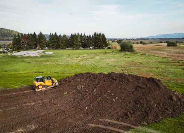 A yellow bulldozer working on a large mound of dirt in a green field, surrounded by trees and a golf course in the background under a clear sky.