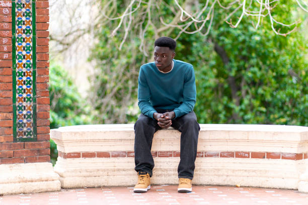 istock Pensive young man sitting on a low wall in a park, looking away 2192557850