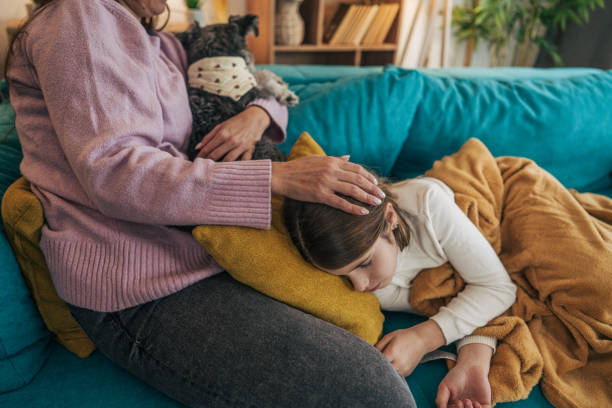 istock Cheerful mom is sitting next to her daughter on the turquoise sofa, using her phone while her daughter lies on the couch, looking unwell. 2192561639