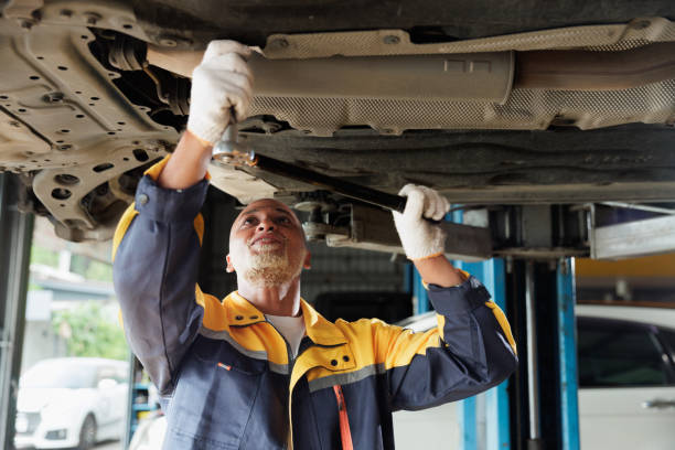 A professional automotive mechanic wearing safety gloves and a uniform is working underneath a raised vehicle in a workshop. He is using a wrench to perform maintenance or repairs on the car's undercarriage. The image highlights technical expertise, precision, and the hands-on nature of automotive repair services in a modern garage environment.