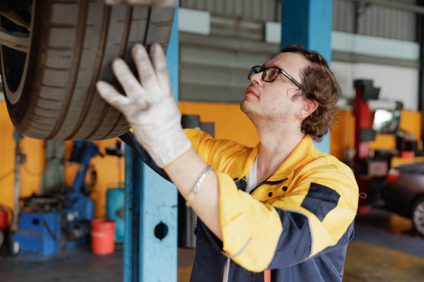 A professional mechanic wearing safety gloves and a uniform is carefully inspecting a car tire elevated on a hydraulic lift inside an auto repair workshop. The workspace features tools, equipment, and vehicles, emphasizing automotive maintenance, safety checks, and repair services.