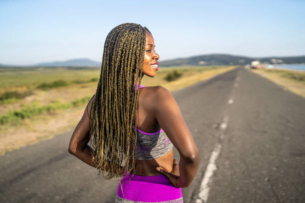 A full length shot of a young woman wearing modern running clothes as she stands on a straight coast road with the diminishing perspective of the highway in front of her and she looks over her shoulder to the side