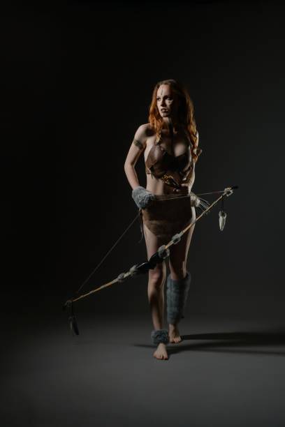 A woman wearing basic tribal clothing holds a handmade bow, exuding strength and confidence in a darkened studio space.