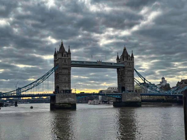 Tower Bridge stretches over the Thames River in London, showcasing its distinct architecture. Dark clouds gather above as evening approaches, creating a moody backdrop to the iconic landmark.