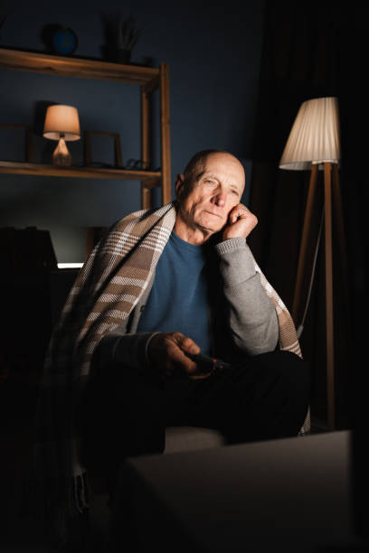 istock Bored pensioner retired wrapped in blanket watching movie alone at home at night using remote control choosing channels with indifferent expression. 2192602024