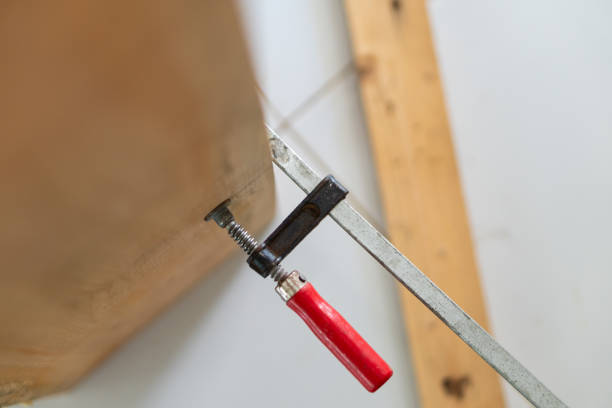 A close-up view of a woodworking clamp with a red handle, securing a wooden piece. The background features wooden beams, emphasizing a workshop environment.