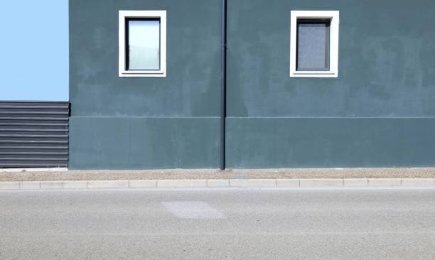 istock Greenish gray plaster wall with gutter in the middle, two windows by sides and black metal fence on left. 2192609896