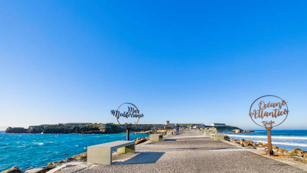 A scenic view of a calm Atlantic ocean with rocky cliffs in the distance, framed by a stone wall and lush green plants in the foreground under a clear blue sky. Camaret sur Mer, Brittany.
