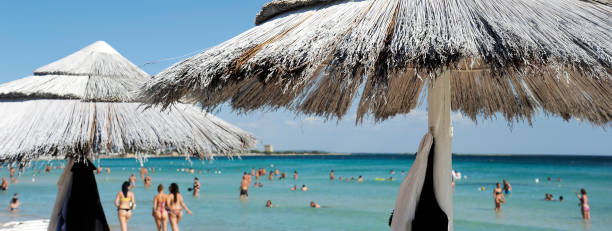 istock Horizontal banner or header with a beautiful beach resort with straw umbrellas on a blue sky and white clouds. On the background bathers  in swimsuits. Beach life and lifestyle concept. 2192615295