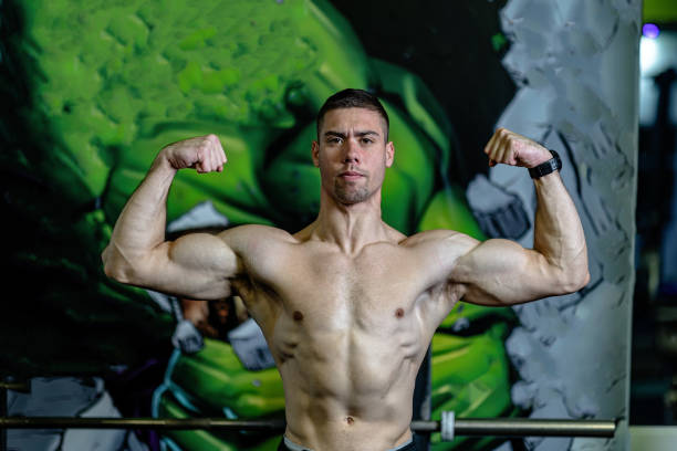 A young man poses in the gym, confidently flexing his biceps to demonstrate his physical fitness and muscular strength. The sporty ambiance highlights the dedication and intensity of achieving a fit physique, evoking feelings of motivation and self-discipline.