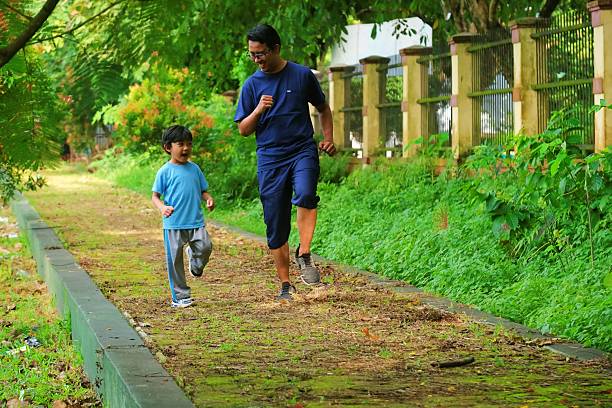 A father and son jogging together on a green pathway surrounded by lush trees. A heartwarming moment of bonding, fitness, and outdoor activity in a serene natural environment.