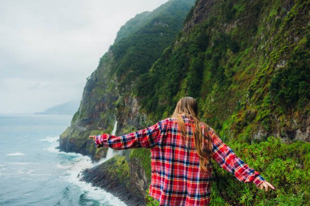 rear view of female with long hair wearing red shirt admiring majestic views of green mountains and waterfall falling into the ocean on Madeira island, Portugal