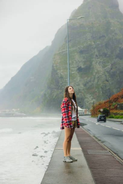 Side view of attractive female traveler in red shirt walking by the road and stormy ocean enjoying the scenic mountain landscape on Madeira island, Portugal