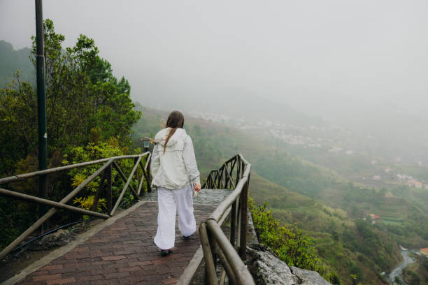 Rear view of female traveler in white clothes enjoying the scenic landscape with lush green mountain and small old towns on Madeira island, Portugal