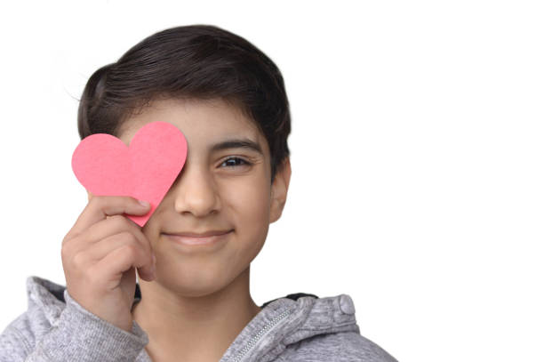 Cut out of one pre teen 12 year old cute out of focus boy holding a paper cutting heart in hand close to his eye covering it, Valentine, related pictures, isolated over white background. There is copy space Horizontal shot of cutout of person in grey jacket holding fun love, valentine, anniversary theme red heart shaped paper cutting close his eye covering it isolated over white background with copy space. Apt for Valentine's Day, Anniversary backdrops, posters, banners, Wedding anniversary, kindness posters, backdrops templates. cute boy show love symbol hand stock pictures, royalty-free photos & images