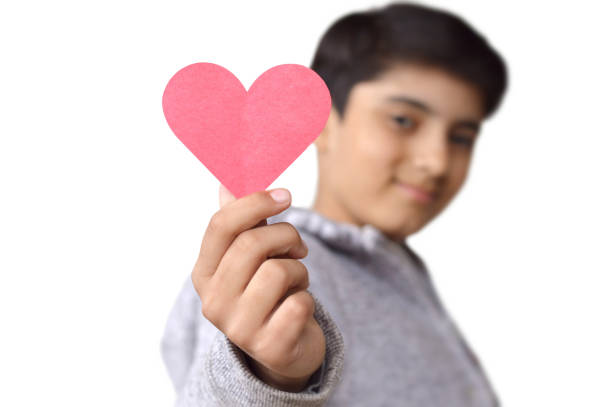 Cut out of one pre teen 12 year old cute out of focus boy holding a paper cutting heart and showing it for love, Valentine, related pictures, isolated over white background. There is depth of field with selective focus on hand and heart Horizontal shot of cutout of person in grey jacket holding fun love, valentine, anniversary theme red heart shaped paper cutting away from his body with one hand isolated over white background with copy space. Apt for Valentine's Day, Anniversary backdrops, posters, banners, Wedding anniversary, care kindness posters, backdrops templates. cute boy show love symbol hand stock pictures, royalty-free photos & images