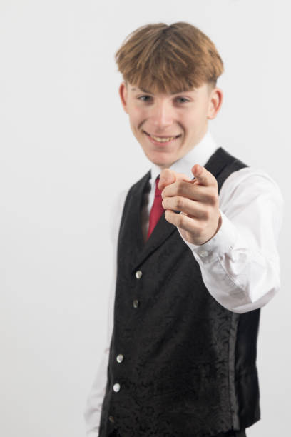 Happy Sixteen year old teenage boy wearing a shirt, tie and waistcoat pointing forward, studio shot against a white background