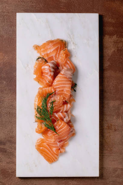Thinly sliced salted salmon fillet served with fresh dill on a white marble board, fine dining. Brown background, top view. Colors of the year 2025.