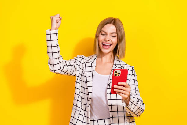 istock Cheerful young woman celebrates success while using smartphone against a vibrant yellow background 2192661351