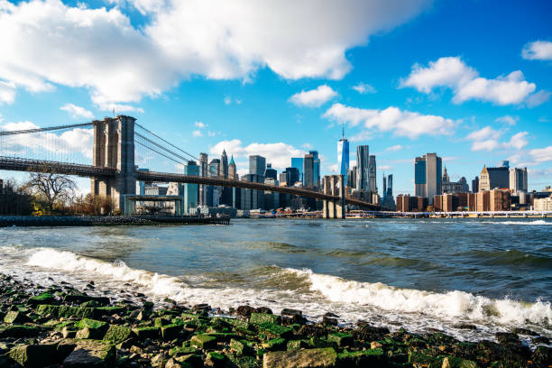 View of the iconic Brooklyn Bridge spanning the East River with New York City's towering Manhattan skyline in the background, captured on a clear day with gentle waves in the foreground.