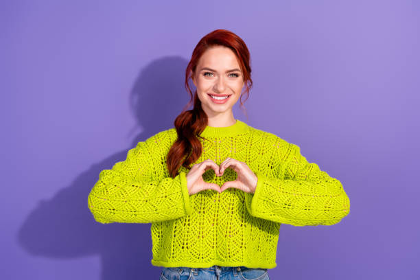 緑のセーターを着た陽気な赤毛の女性が、鮮やかな紫色の背景にハートのジェスチャーをしています - 自分 ストックフォトと画像