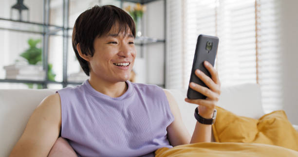 istock Gen Z queer man non-binary sitting easy relax happy read news on web page app at home sofa. Asia people LGBT young adult guy play web3, store shop e-commerce buyer, virtual retail network cyber paying 2192664073