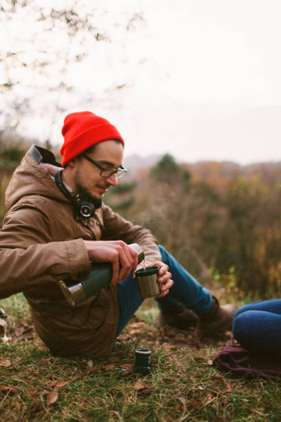 Green thermos with delicious hot tea on old mossy tree in focus. The background of the autumn forest is blurred.