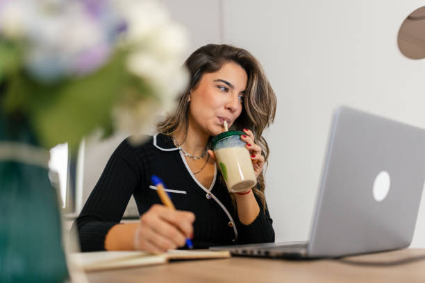 Beautiful woman sitting at the table, drinking protein smoothie shake, working on her laptop and writing something in her notebook.