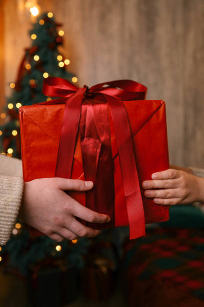 istock Hands exchanging a festive gift box with a red ribbon under a beautifully decorated Christmas tree. Cozy holiday setting with candles, miniature houses, and a warm atmosphere 2192676231