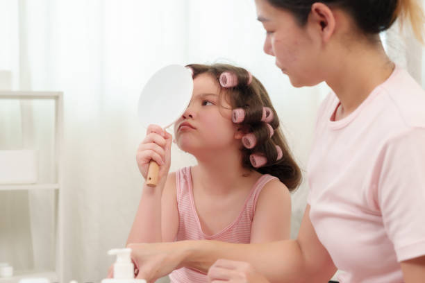 Asian single mother teaching young daughter makeup and hairstyling using mirror and rollers in cozy home environment. Bonding moment showcasing family love, creativity, and beauty exploration.