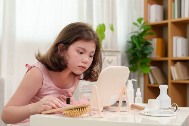 istock Young girl leaning forward at table while carefully observing her reflection in mirror during beauty session at home. Scene depicting focus, curiosity, and self-expression in relaxed setting. 2192676897