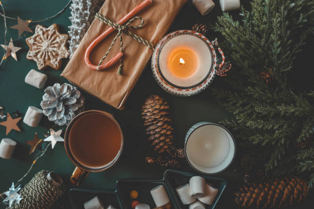 istock Cozy Christmas setup featuring a wrapped gift with a candy cane, a lit candle, tea, an open book, pinecones, marshmallows, and evergreen branches on a green background 2192676915