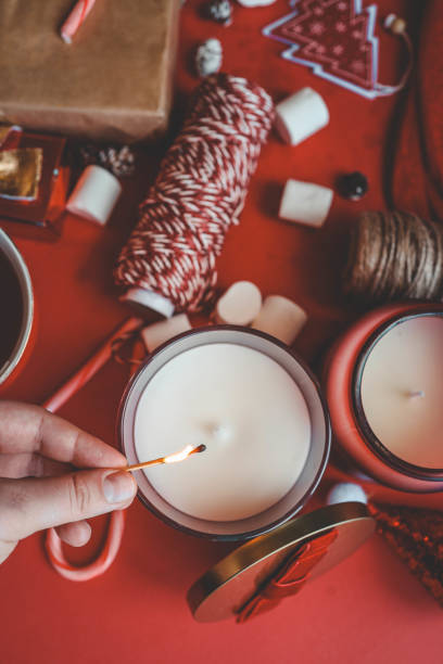 istock Hand lighting a white candle surrounded by holiday decorations on a red background. Festive mood with candy canes, marshmallows, gift boxes, and twine, creating a warm Christmas atmosphere 2192676930