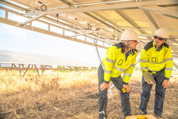 istock Two solar technicians in bright reflective jackets and helmets are inspecting the wiring under solar panels. One holds laptop for data logging, while the other examines electrical connections closely. 2192688769