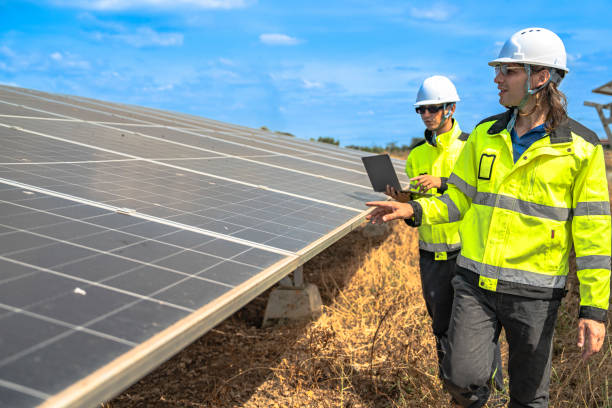 istock Two technicians in safety gear and reflective jackets inspect a solar panel installation at a solar farm. They are engaged in maintenance and operational checks under a clear blue sky. 2192689605