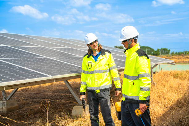 istock Two engineers wearing safety helmets and reflective jackets inspect a solar panel installation. One uses a drill tool, while the other checks the panel surface under a bright, sunny sky. 2192689649