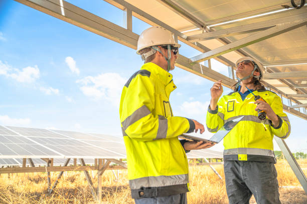 istock Two solar technicians in bright reflective jackets and helmets are inspecting the wiring under solar panels. One holds laptop for data logging, while the other examines electrical connections closely. 2192690329