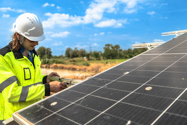 istock A solar technician in a high-visibility jacket and helmet stands next to a solar panel, with a serene landscape featuring a pond and distant greenery under a partly cloudy sky. 2192690504