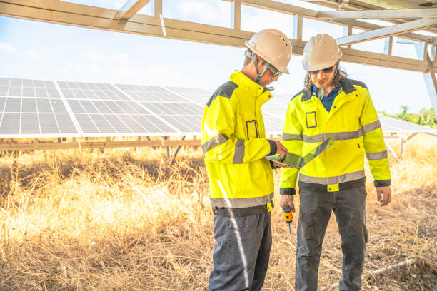 istock Two solar technicians in bright reflective jackets and helmets are inspecting the wiring under solar panels. One holds laptop for data logging, while the other examines electrical connections closely. 2192691373