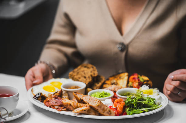 istock A hearty breakfast plate featuring eggs, bacon, sausages, fresh vegetables, and side dips, served with slices of bread. A cozy dining scene with a warm beverage nearby. 2192697151