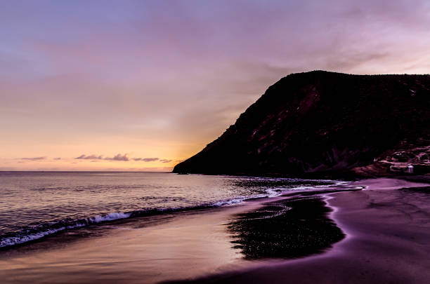 A beautiful sunset over the ocean with a mountain in the background. The sky is a mix of purple and orange hues, creating a serene and peaceful atmosphere. The water is calm