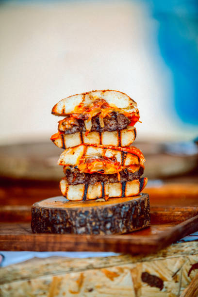 Close-up of gourmet burger halves stacked neatly on a rustic wooden base. The burgers showcase perfectly grilled buns with visible char lines, juicy beef patties, melted cheese, and rich sauce dripping from the layers. A blurred background enhances the focus on the intricate details of the burger, making it an ideal visual for artisanal food and gourmet dining promotions