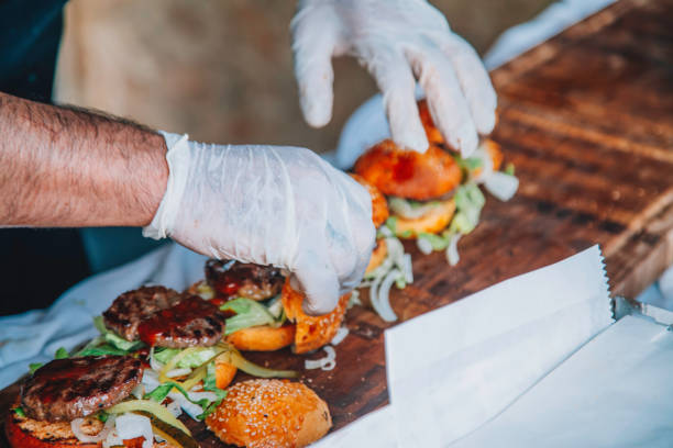 A close-up view of a chef's hands assembling gourmet burgers on a rustic wooden board. The chef is wearing white gloves for hygiene, carefully placing fresh ingredients such as lettuce, onion, and juicy burger patties onto sesame seed buns. The scene captures the artistry and precision in crafting delicious, high-quality burgers, highlighting the fresh produce and attention to detail in a professional kitchen setting
