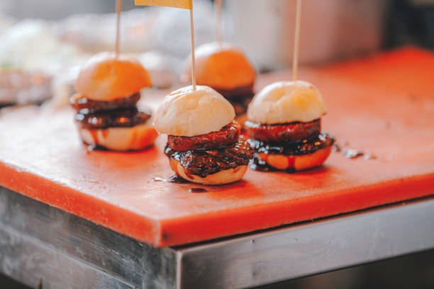 A close-up view of mini slider burgers neatly arranged on a wooden surface. The sliders feature soft, freshly baked buns and juicy beef patties, coated in a rich, dark glaze. Each burger is secured with a skewer for easy handling. The smooth texture of the buns contrasts with the glossy sauce, creating a mouthwatering visual ideal for catering or event menus