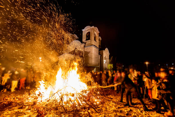 A vibrant nighttime gathering of people near a bright bonfire in front of a church. The sparks illuminate the festive atmosphere as individuals partake in a traditional celebration, uniting the community. The scene highlights a warm and joyous event filled with cultural and spiritual significance, set against dark, contrasting skies.