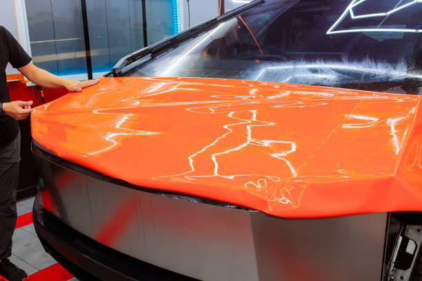 Bright orange vinyl wrap application on a car's hood at a vehicle customization workshop in the afternoon